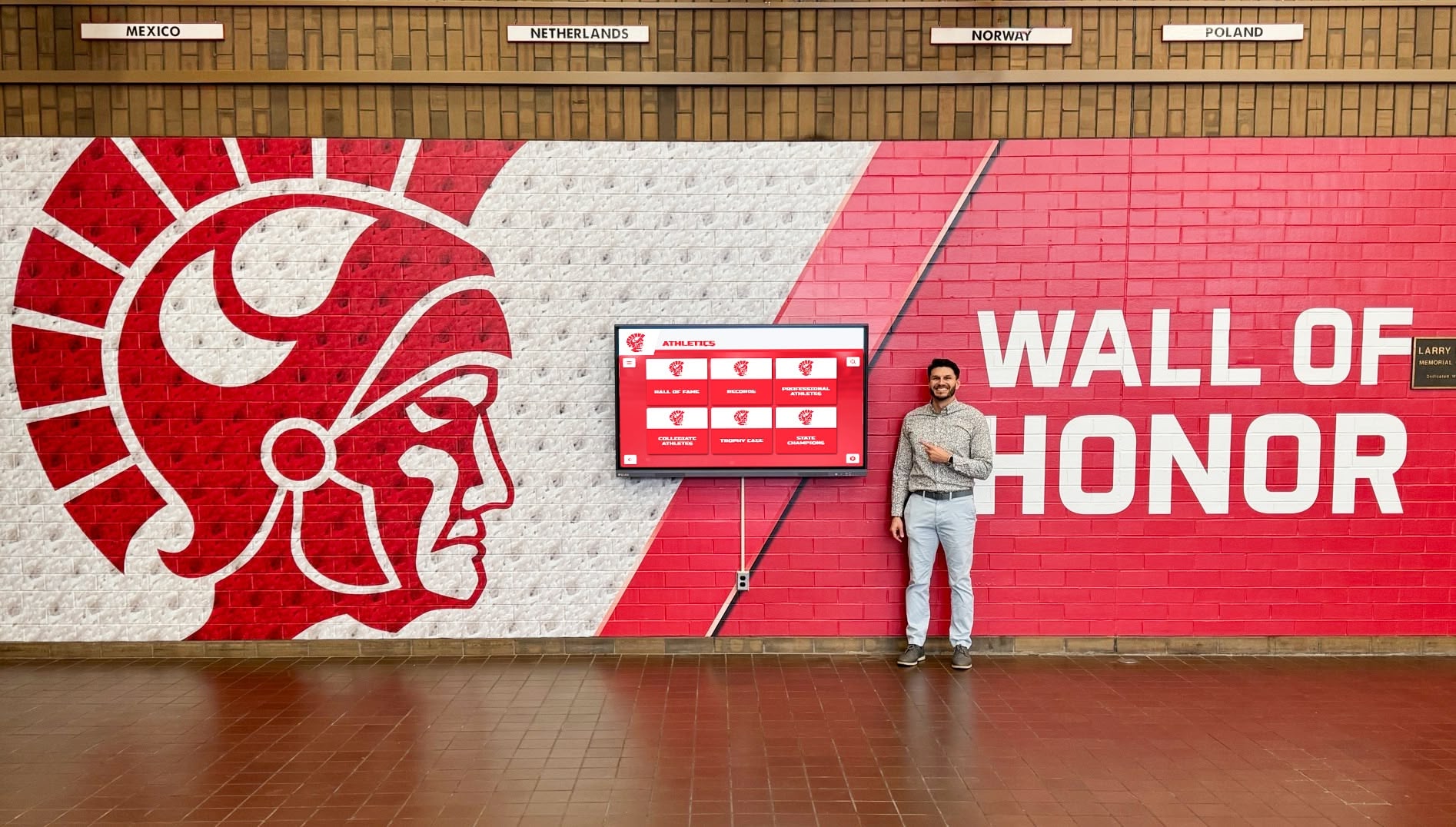 man pointing at red trojan wall of honor in school hallway